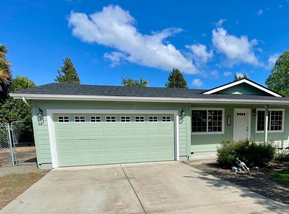 Single-story green house with a garage, driveway, and bright blue sky in the background.