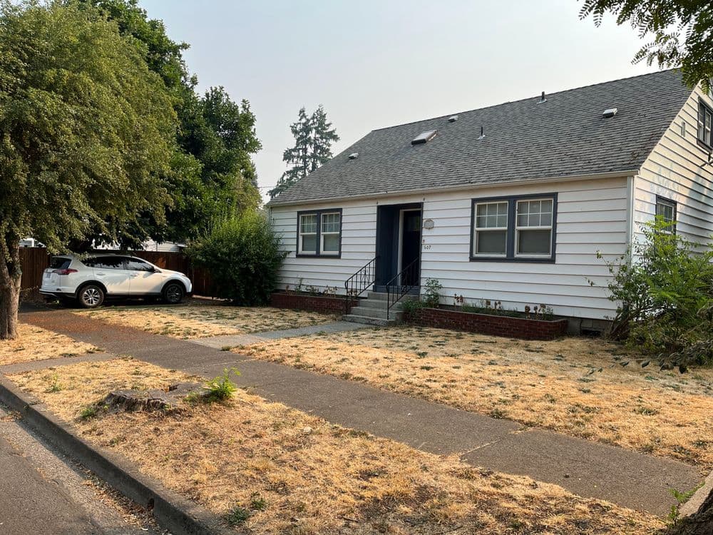 White single-story house with a gray roof, parked car, and dry lawn in suburban neighborhood.