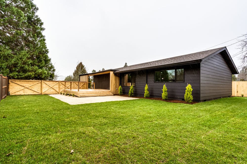 Modern black house with wooden deck and fenced yard, surrounded by lush green grass.