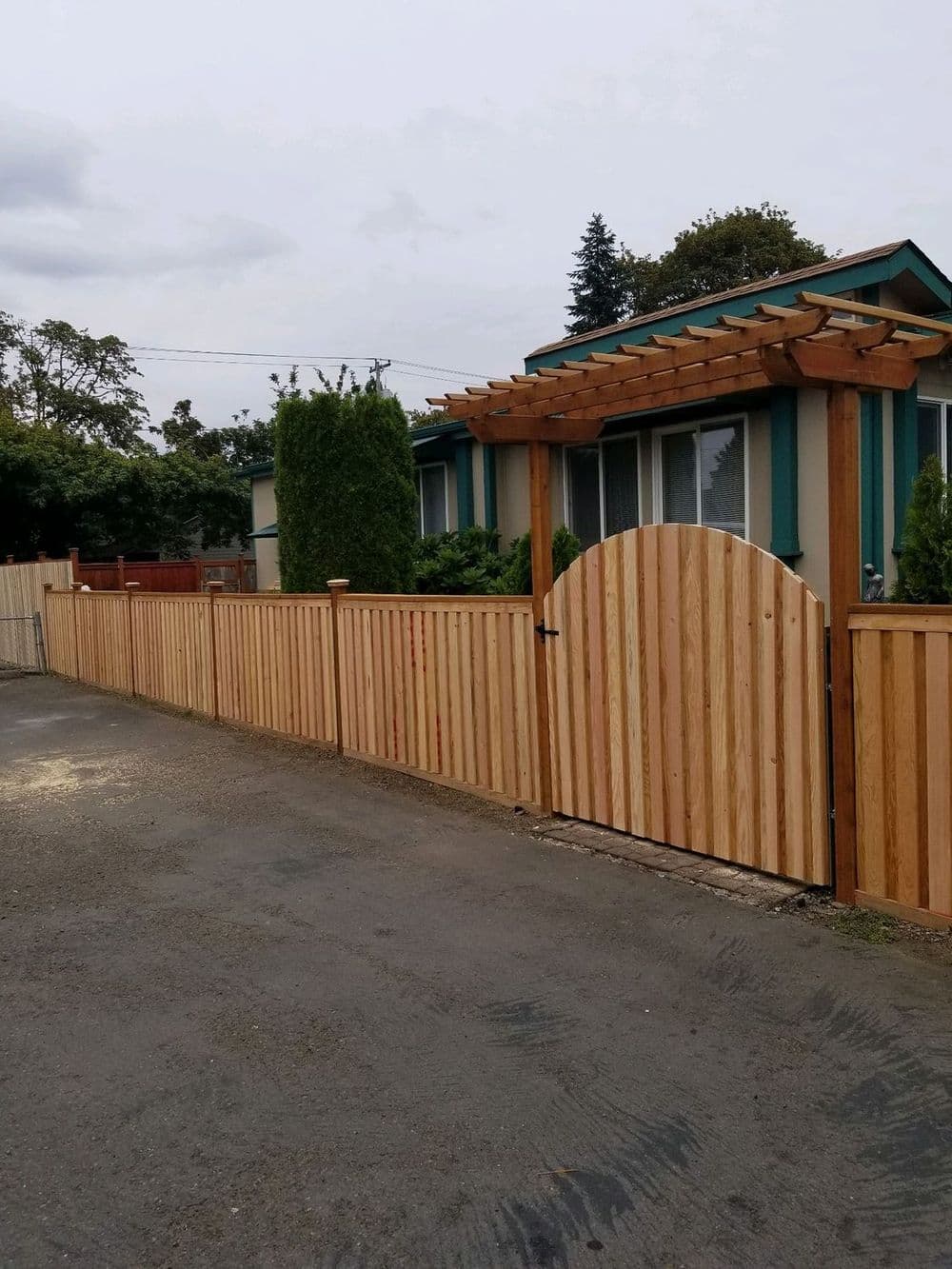 Wooden fencing with an arched gate, leading to a house surrounded by greenery.