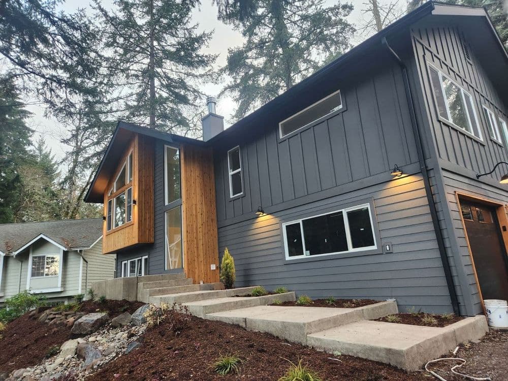Modern two-story house with wood and gray siding, surrounded by tall trees and landscaping.