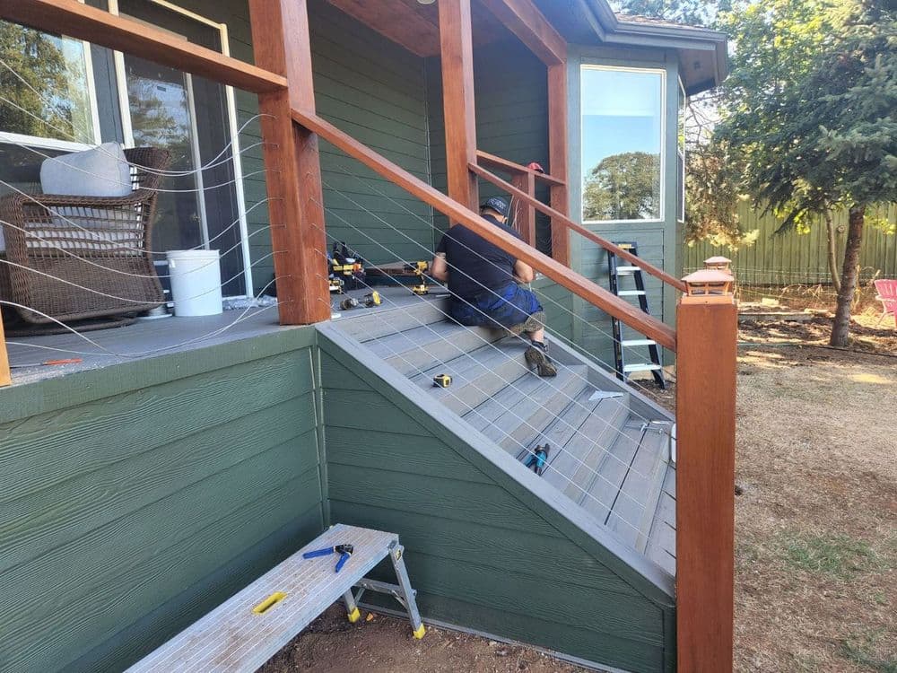 Man installing cable railing on outdoor staircase, surrounded by tools and a green house.